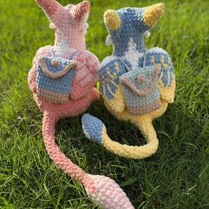 May include: Two handmade crochet plush toys in pink and blue, each with a small backpack. The toys have long tails and are set on a bed of green grass. The text "PATTERN!" is at the top of the image.