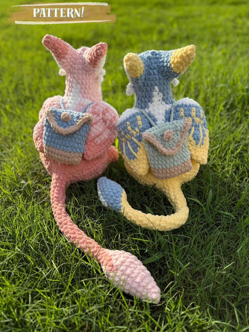 May include: Two handmade crochet plush toys in pink and blue, each with a small backpack. The toys have long tails and are set on a bed of green grass. The text "PATTERN!" is at the top of the image.