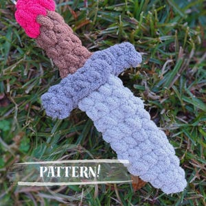 May include: A handmade crochet sword in shades of pink, brown, and gray, lying on a bed of green grass. The word "PATTERN!" is displayed across the image.