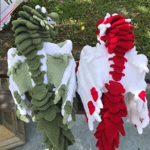 May include: Two crocheted dragon plushies, one green and one red, with white wings. The dragons are sitting on a bench with a grassy background.
