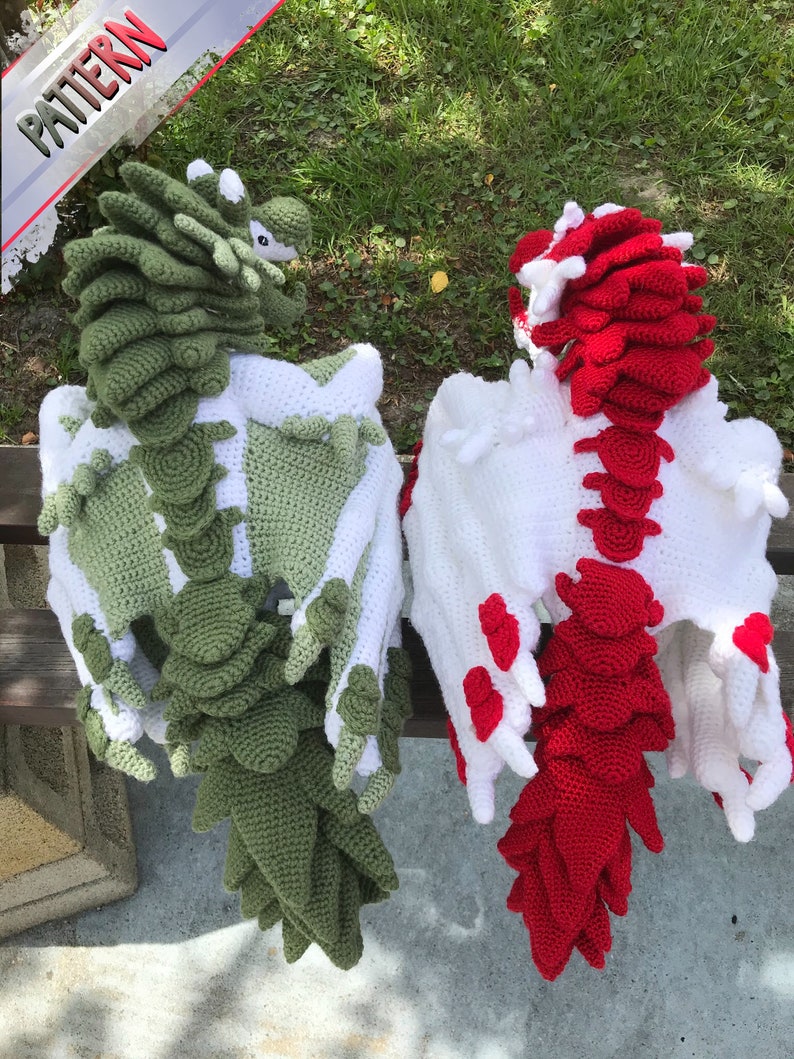 May include: Two crocheted dragon plushies, one green and one red, with white wings. The dragons are sitting on a bench with a grassy background.