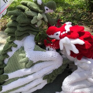 May include: Two crocheted dragon plushies, one green and white, the other red and white, are posed together on a grassy surface.