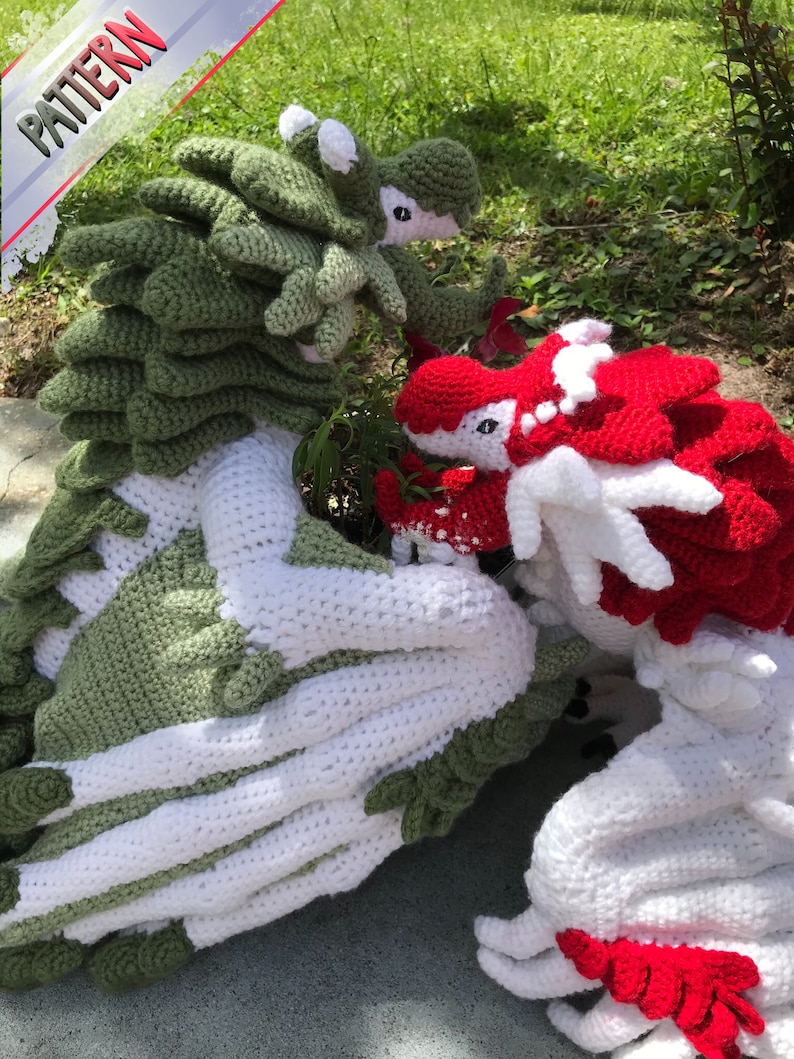 May include: Two crocheted dragon plushies, one green and white, the other red and white, are posed together on a grassy surface.