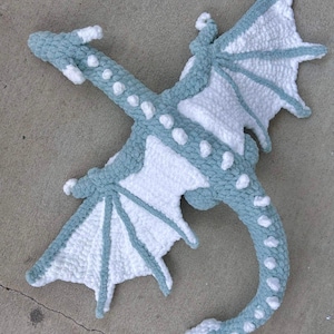 May include: A crocheted dragon plush toy with a light blue and white color scheme. The dragon is lying on its back with its wings spread out.