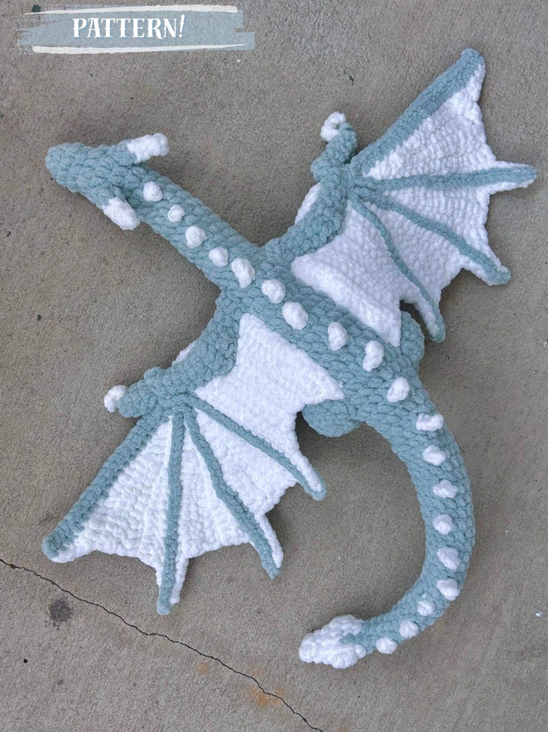 May include: A crocheted dragon plush toy with a light blue and white color scheme. The dragon is lying on its back with its wings spread out.