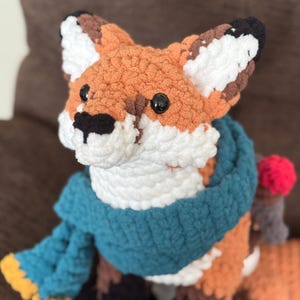 K&ouml;nnte beinhalten: Eine handgefertigte H&auml;kelfuchs-Puppe mit orangefarbenen, wei&szlig;en, braunen und schwarzen Details. Der Fuchs tr&auml;gt einen blauen Schal und hat schwarze Knopfaugen. Das Wort "PATTERN!" ist unten im Bild aufgedruckt.