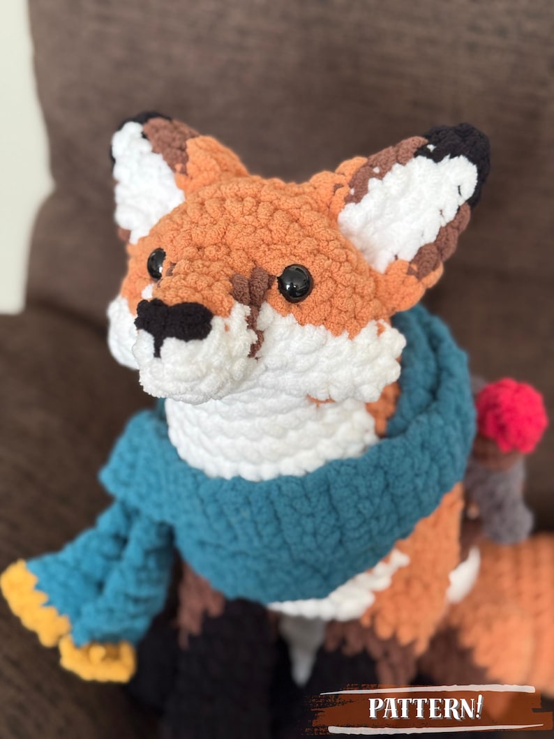 K&ouml;nnte beinhalten: Eine handgefertigte H&auml;kelfuchs-Puppe mit orangefarbenen, wei&szlig;en, braunen und schwarzen Details. Der Fuchs tr&auml;gt einen blauen Schal und hat schwarze Knopfaugen. Das Wort "PATTERN!" ist unten im Bild aufgedruckt.