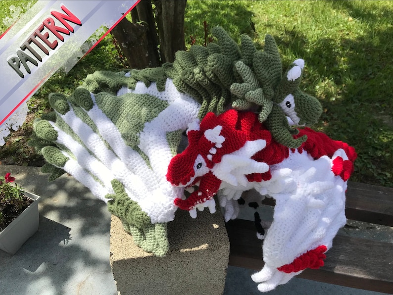 May include: Three crocheted dragons, one green, one white, and one red, are stacked on top of each other. The dragons are all facing the same direction and have their mouths open.
