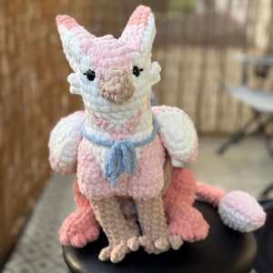 May include: A handmade crochet plush toy in the shape of a mythical creature, mainly pink and white. The toy has a light blue scarf and a tail. The word "PATTERN!" is written on a banner above the toy.