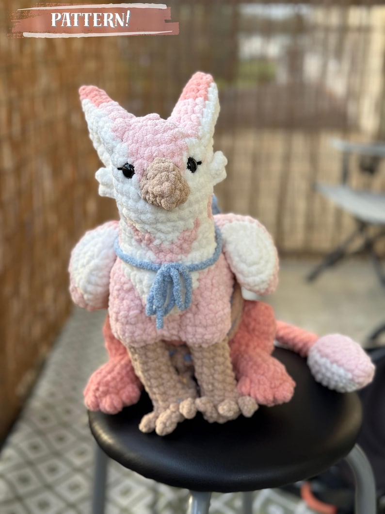 May include: A handmade crochet plush toy in the shape of a mythical creature, mainly pink and white. The toy has a light blue scarf and a tail. The word "PATTERN!" is written on a banner above the toy.