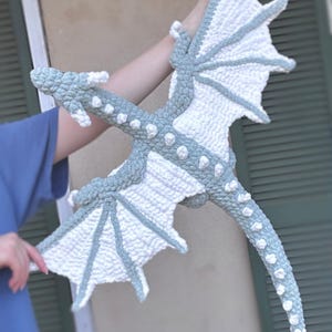 May include: A crocheted dragon plush toy with white and gray wings and a long tail. The dragon is made with a textured yarn and has a whimsical, friendly expression.