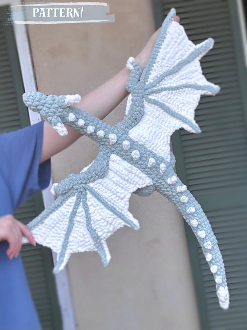 May include: A crocheted dragon plush toy with white and gray wings and a long tail. The dragon is made with a textured yarn and has a whimsical, friendly expression.