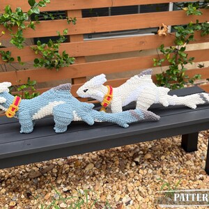 May include: Two crocheted dragon toys, one light blue and one white, rest on a black bench. The blue dragon has a white belly and a yellow and red collar. The white dragon has a gray tail and collar. The word "PATTERN!" is in the bottom right corner.