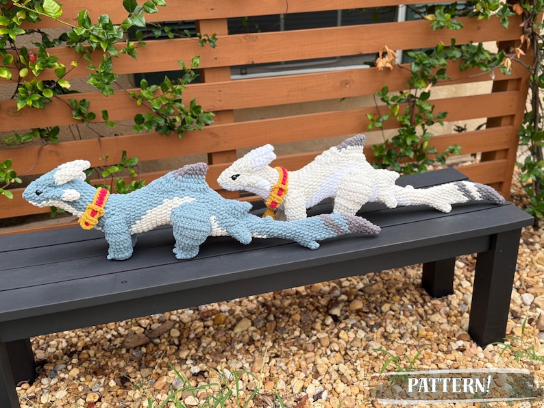 May include: Two crocheted dragon toys, one light blue and one white, rest on a black bench. The blue dragon has a white belly and a yellow and red collar. The white dragon has a gray tail and collar. The word "PATTERN!" is in the bottom right corner.