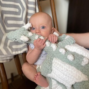 May include: A baby is sitting in a chair holding a green and white crocheted dragon blanket. The blanket has a textured, fluffy look.
