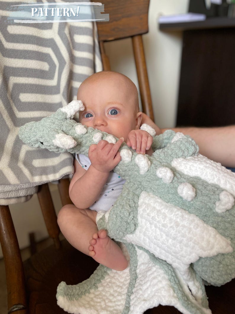 May include: A baby is sitting in a chair holding a green and white crocheted dragon blanket. The blanket has a textured, fluffy look.