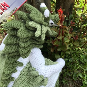 May include: Green and white crocheted dragon amigurumi toy with a spiky back and a white face. The toy is sitting in a garden setting.