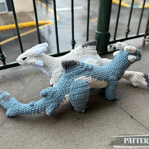 May include: Three crocheted dragon toys in shades of blue, white, and gray. The dragons have detailed features and are displayed on a concrete surface. The word "PATTERN!" is printed on the bottom of the image.