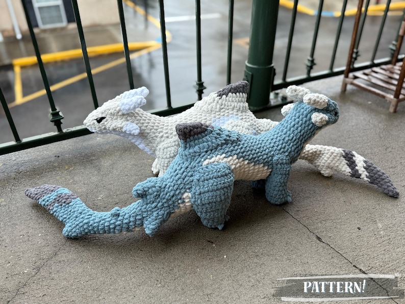 May include: Three crocheted dragon toys in shades of blue, white, and gray. The dragons have detailed features and are displayed on a concrete surface. The word "PATTERN!" is printed on the bottom of the image.