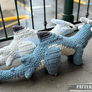 May include: Three crocheted dinosaur plushies in shades of blue and white, with gray accents. The dinosaurs are positioned on a gray surface. The word "PATTERN!" is visible in the image.