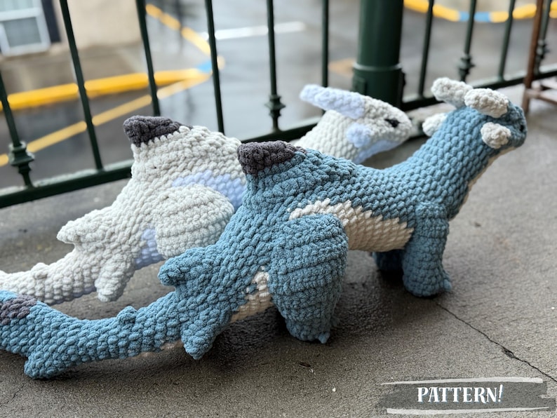 May include: Three crocheted dinosaur plushies in shades of blue and white, with gray accents. The dinosaurs are positioned on a gray surface. The word "PATTERN!" is visible in the image.