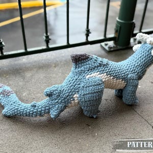 May include: A handmade blue crochet dinosaur toy with white and gray accents. The dinosaur has a long tail and is lying on a concrete surface. The word "PATTERN!" is visible in the image.