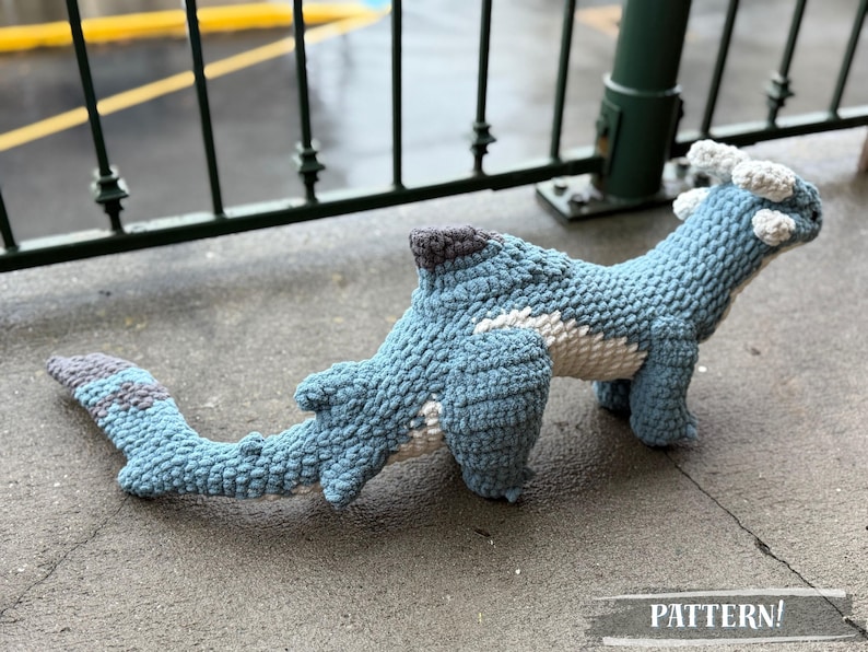 May include: A handmade blue crochet dinosaur toy with white and gray accents. The dinosaur has a long tail and is lying on a concrete surface. The word "PATTERN!" is visible in the image.