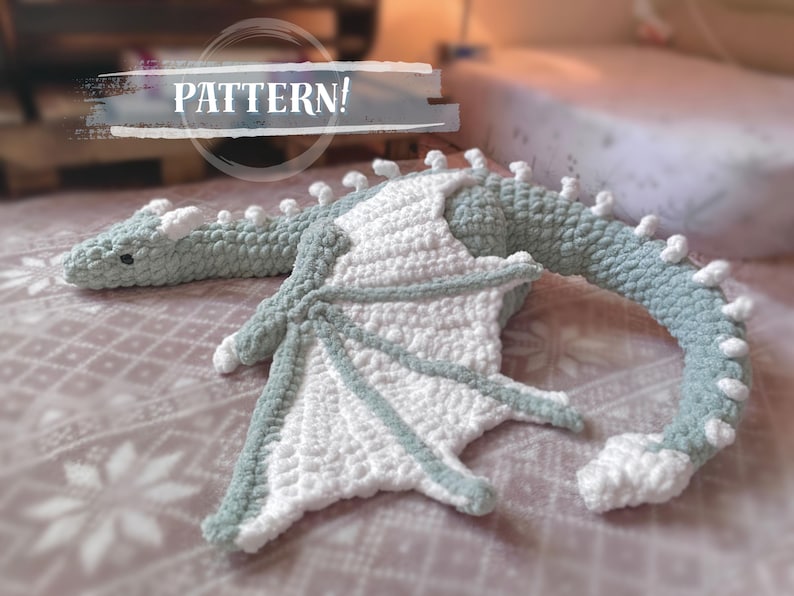 May include: A crocheted dragon plush toy with a light green body and white wings. The dragon is lying on a white and gray patterned surface. The text "PATTERN!" is visible in the background.
