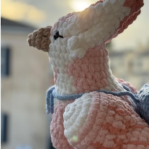 May include: A handmade crochet dinosaur in shades of pink, white, and brown. The plush toy has a textured, fluffy appearance with a blue scarf. The word "PATTERN!" is written in a pink banner at the top.