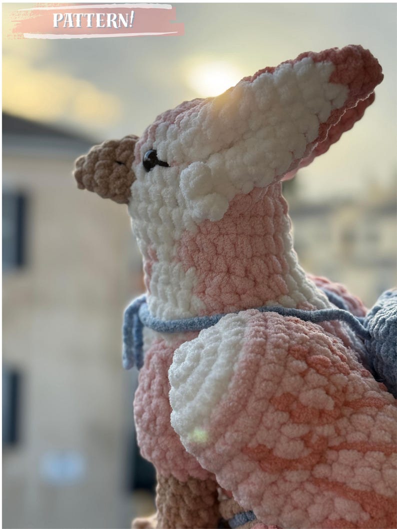 May include: A handmade crochet dinosaur in shades of pink, white, and brown. The plush toy has a textured, fluffy appearance with a blue scarf. The word "PATTERN!" is written in a pink banner at the top.