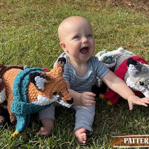 K&ouml;nnte beinhalten: Ein Baby spielt mit drei geh&auml;kelten Tierkissen: einem Fuchs mit t&uuml;rkisfarbenem Schal, einem Reh mit Blumenkranz und einem grauen Wolf mit rotem Schal. Die Kissen liegen auf gr&uuml;nem Gras.