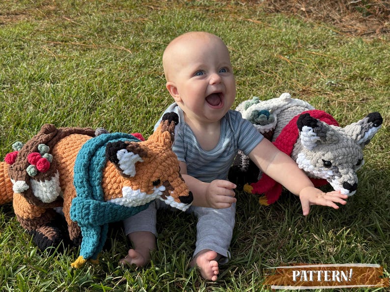 K&ouml;nnte beinhalten: Ein Baby spielt mit drei geh&auml;kelten Tierkissen: einem Fuchs mit t&uuml;rkisfarbenem Schal, einem Reh mit Blumenkranz und einem grauen Wolf mit rotem Schal. Die Kissen liegen auf gr&uuml;nem Gras.