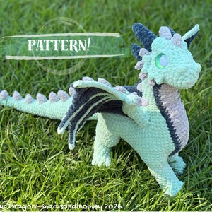 May include: A crocheted dragon toy in mint green, dark blue, and light gray yarn. The creature features a long tail with spikes, wings, and glowing green eyes. Text on the image reads: PATTERN! The Archaic Dragon - madisondinomay 2026.
