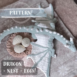 Może przedstawiać: Szydełkowy smok z gniazdem i jajkami. Smok jest jasnoniebieski i biały, z teksturowanym ciałem i skrzydłami. Gniazdo jest brązowe, a jajka białe. Obraz zawiera tekst "PATTERN!" i "DRAGON + NEST + EGGS!"