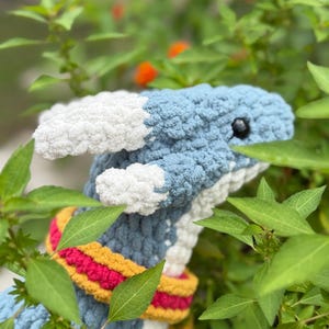 May include: A handmade crochet dragon with blue, white, and yellow accents. The dragon has a blue body, white accents, and a colorful scarf. The text "PATTERN!" is visible in the upper left corner. The dragon is surrounded by green foliage.