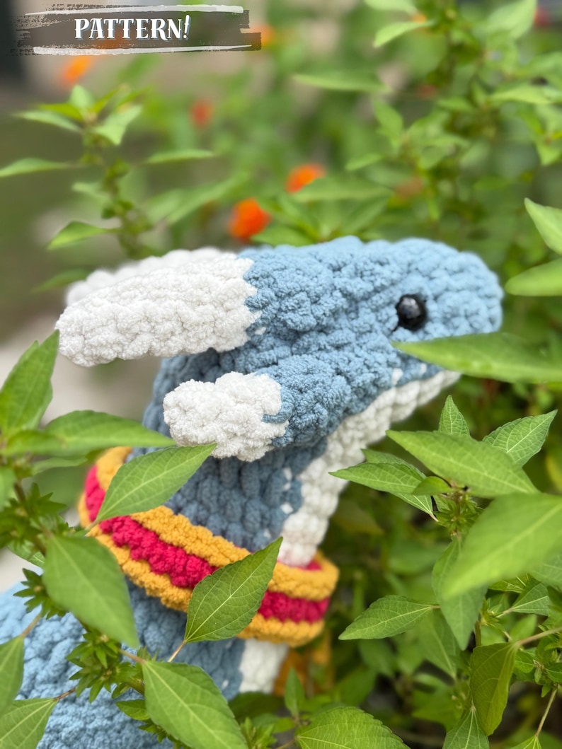 May include: A handmade crochet dragon with blue, white, and yellow accents. The dragon has a blue body, white accents, and a colorful scarf. The text "PATTERN!" is visible in the upper left corner. The dragon is surrounded by green foliage.