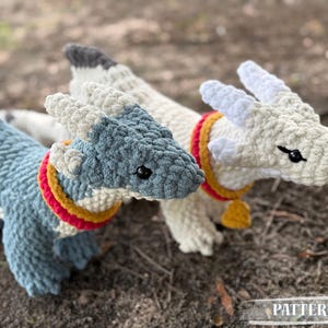 May include: Three handmade crochet dragons in shades of blue and white, with red and yellow collars. The dragons have black eyes and are lying on a bed of dirt. The word "PATTERN!" is at the bottom of the image.