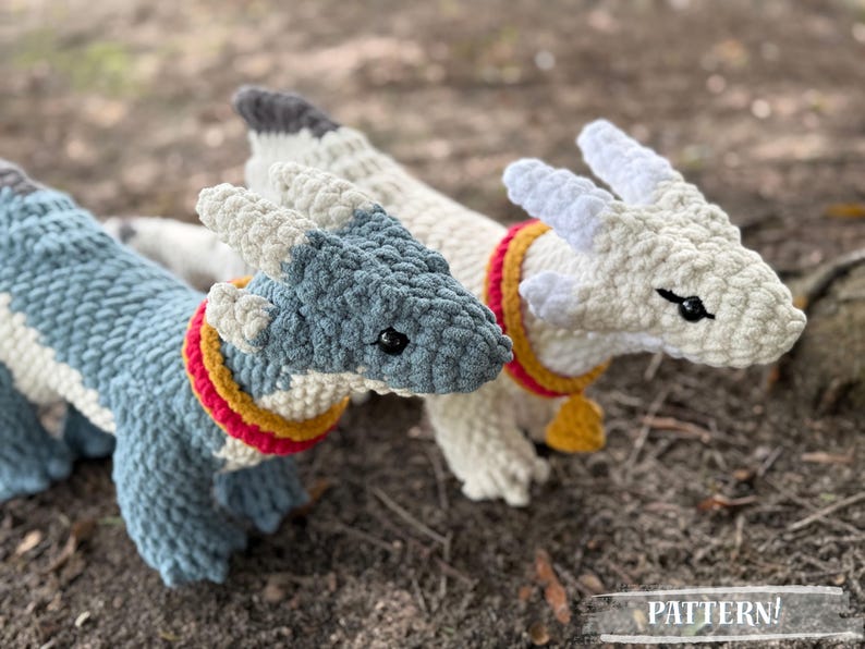 May include: Three handmade crochet dragons in shades of blue and white, with red and yellow collars. The dragons have black eyes and are lying on a bed of dirt. The word "PATTERN!" is at the bottom of the image.