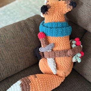 K&ouml;nnte beinhalten: Geh&auml;keltes Fuchspl&uuml;schtier in Orange, Braun und Wei&szlig;, mit blauem Schal, Schwert und Beerenb&uuml;ndel. Der Text "PATTERN!" ist sichtbar. Der Fuchs sitzt auf einem braunen Sofa.