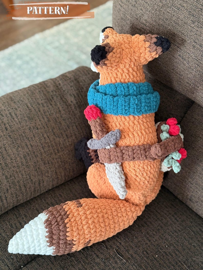 K&ouml;nnte beinhalten: Geh&auml;keltes Fuchspl&uuml;schtier in Orange, Braun und Wei&szlig;, mit blauem Schal, Schwert und Beerenb&uuml;ndel. Der Text "PATTERN!" ist sichtbar. Der Fuchs sitzt auf einem braunen Sofa.