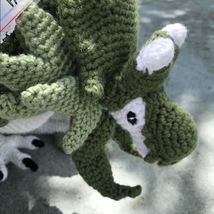 May include: Close-up of a green crocheted dragon with white accents. The dragon has a large head with a pointed snout and a single black eye. The dragon's body is covered in green crochet stitches.