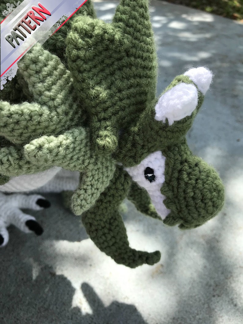 May include: Close-up of a green crocheted dragon with white accents. The dragon has a large head with a pointed snout and a single black eye. The dragon's body is covered in green crochet stitches.