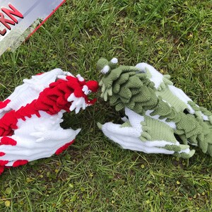 May include: Two crocheted dragons, one red and white and one green and white, are lying on a bed of green grass.