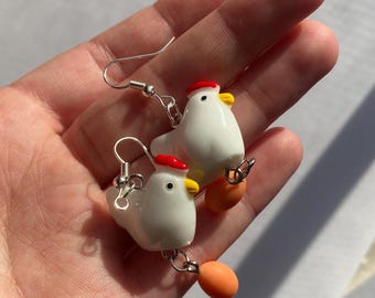 Pode incluir: Um par de brincos de novidade com galinhas de cerâmica branca com bonés vermelhos e bicos amarelos. Cada galinha tem um ovo laranja pendurado por baixo. Os brincos são suspensos por ganchos de orelha prateados.