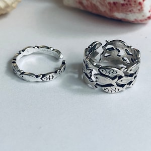 May include: Two silver rings featuring a fish design. The ring on the left is a simple band with fish outlines. The ring on the right is a stack of fish, each with a detailed design.