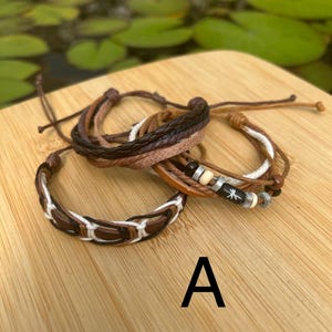 May include: A set of three layered bracelets featuring brown leather cords, braided dark brown leather, and white cord accents. One bracelet includes small cylindrical beads in black, white, and metallic silver, with a white star-like symbol.