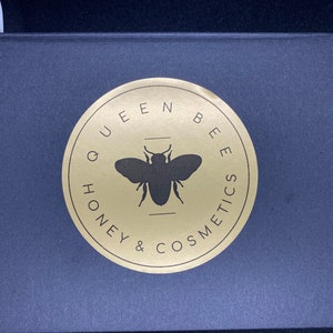 May include: A black box with a gold circle logo that says "Queen Bee Honey & Cosmetics" with a silhouette of a bee in the center.