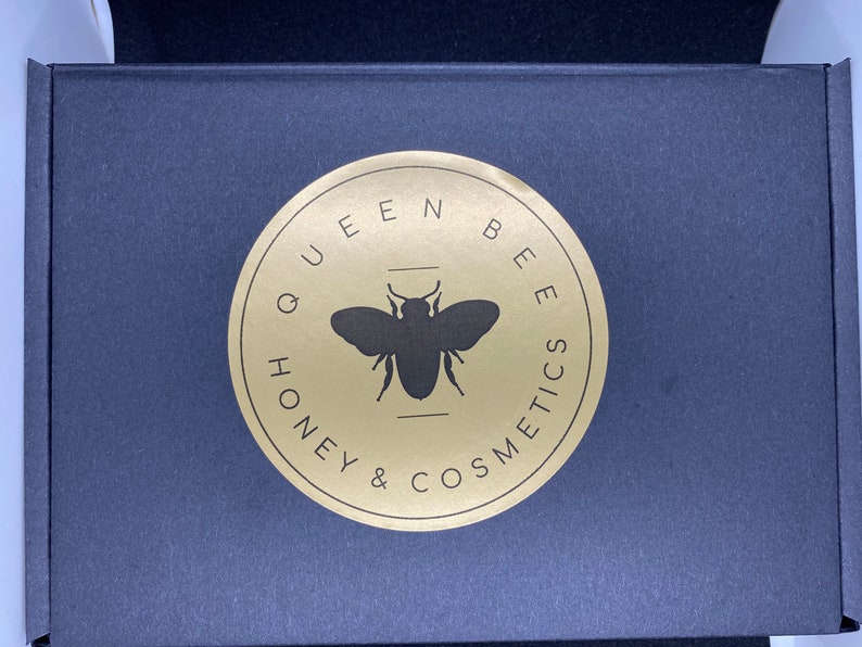 May include: A black box with a gold circle logo that says "Queen Bee Honey & Cosmetics" with a silhouette of a bee in the center.