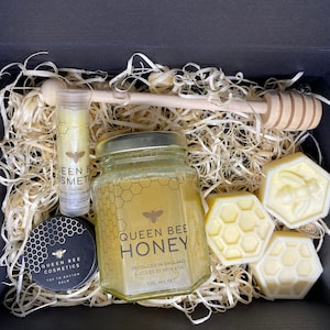 May include: A gift box filled with honey-themed bath and body products. The box contains a jar of honey, a honey dipper, a lip balm, and three honey-shaped soaps. The products are all labeled with the words "Queen Bee Cosmetics" and "Queen Bee Honey".