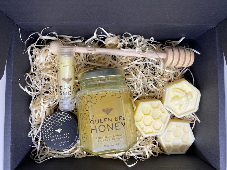 May include: A gift box filled with honey-themed bath and body products. The box contains a jar of honey, a honey dipper, a lip balm, and three honey-shaped soaps. The products are all labeled with the words "Queen Bee Cosmetics" and "Queen Bee Honey".